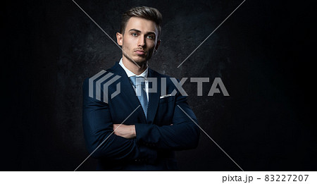 Conceptual portrait of a young, stylish business man, in a blue suit, hands on a cross, against a dark textured background. 83227207