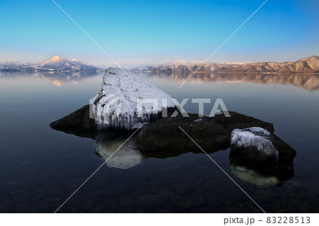 雪と氷をかぶった岩と猪苗代湖に姿を映す夕日を浴びた雪の磐梯山 83228513