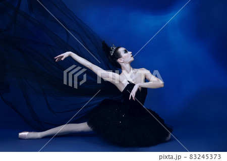 Portrait of adorable female ballet dancer in black tutu and stage make-up sitting on floor isolated on blue studio backgorund 83245373