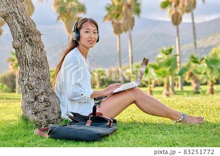 Young woman in headphones with laptop sitting on the grass in the park Young woman in headphones with laptop sitting on the grass in the park 83251772