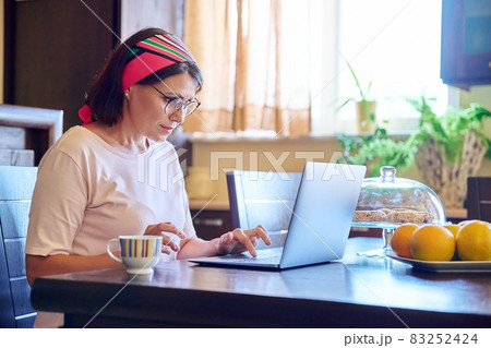 Middle-aged woman sitting at home in the kitchen with a laptop and a cup of coffee 83252424