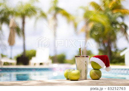 Snowman made of apples in a Christmas hat standing near swimming pool with a cocktail on the palm trees background. Selective focus, blurred background. 83252763