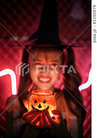 Charming witch holding halloween pumpkin candle cup in red light on dark background Charming witch holding halloween pumpkin candle cup in red light on dark background 83260676