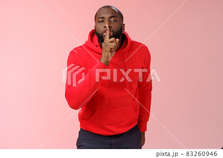 Shhh it taboo. Portrait sensual funny cute african-american bearded man in red hoodie bending camera show shush gesture index finger on lips telling secret asking keep quiet not tell anyone mystery 83260946