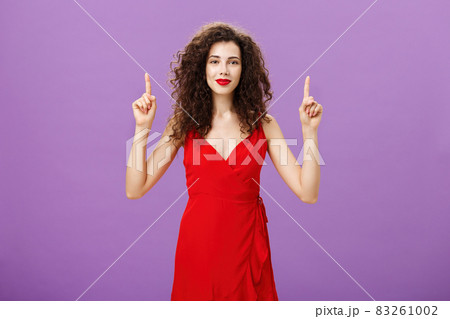 Feeling on top after making stylish makeup. Cute and tender glamourous european female in red evening dress and lipstick with dark curly hair smiling gently and pointing up with raised hands 83261002