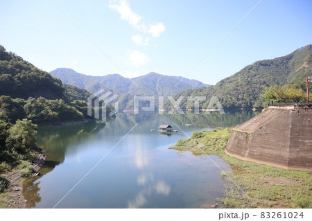 青空と雲と白む空と周囲を写す水鏡の湖面の山に囲まれた小屋の浮かぶ湖とコンクリートのダムのある風景 青空と雲と白む空と周囲を写す水鏡の湖面の山に囲まれた小屋の浮かぶ湖とコンクリートのダムのある風景 83261024