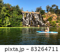 The girl swims on sup near the waterfall. Rest on the river. A group of people near an artificial waterfall. Green trees around the waterfall, blue sky with no clouds 83264711