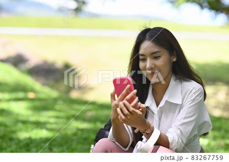 Smiling young asian woman using smart phone while sitting in the park. 83267759