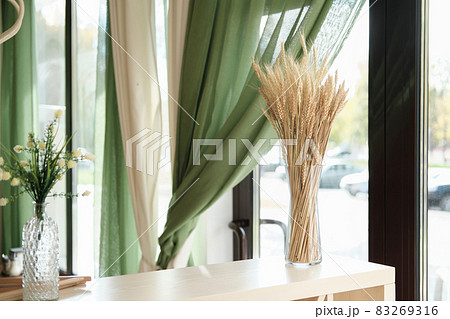 partial view of stylish interior of cafe or terrace with natural decor. ear of wheat in a glass vase on light wooden table, green curtains and flowers. window view to a street. 83269316