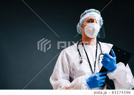 Young man nurse hospital worker in medical protective mask, gloves and protective wear isolated on dark background with copy space. 83274396