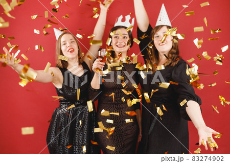 Three delighted women in black dress and party hats over red background with golden confetti. Female party 83274902