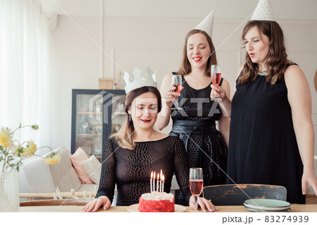 Three women friends celebrating 30s birthday together gathering at table at home with wine and birthday cake with candles. Women in party hat and birthday girl in crown blows out candles. Three women friends celebrating 30s birthday together gathering at table at home with wine and birthday cake with candles. Women in party hat and birthday girl in crown blows out candles. 83274939