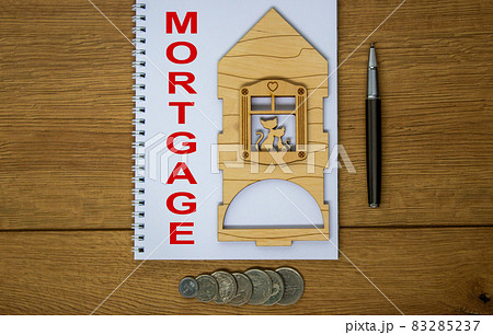 White note with inscription 'mortgage' on beautiful wooden table, metalic pen, coins and miniature wooden house. Business concept. 83285237