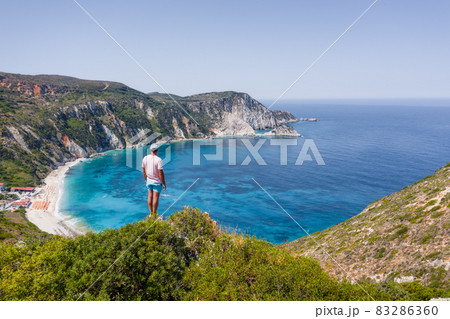 Man standing on top of a rock with an exciting feeling of freedom, looking at Petani Beach. Kefalonia ionian island, Greece Man standing on top of a rock with an exciting feeling of freedom, looking at Petani Beach. Kefalonia ionian island, Greece 83286360