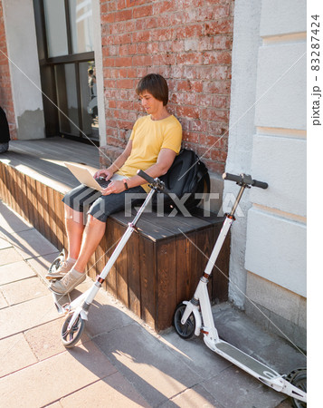 Man sits with laptop and kick scooter in street cafe. Freelancer at work. Student learns remotely from outdoors. Modern lifestyle. Summer vibes. Open air workplace. Man sits with laptop and kick scooter in street cafe. Freelancer at work. Student learns remotely from outdoors. Modern lifestyle. Summer vibes. Open air workplace. 83287424