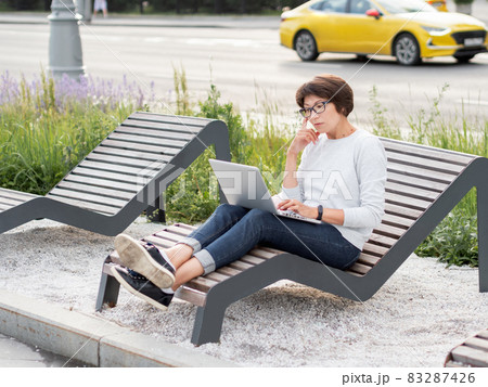 Woman sits with laptop on urban park bench. Freelancer at work. Student learns remotely from outdoors. Modern lifestyle. 83287426