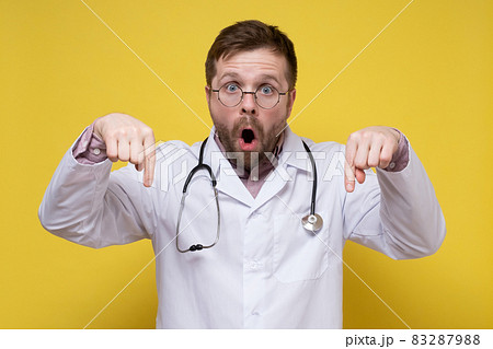 Agitated doctor in a white coat with a stethoscope around neck points down with index fingers, looks 83287988