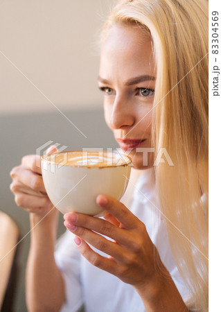 Close-up face of attractive young blonde woman drinking coffee hot cappuccino coffee with beautiful pattern sitting in cafe. Close-up face of attractive young blonde woman drinking coffee hot cappuccino coffee with beautiful pattern sitting in cafe. 83304569