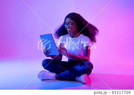 Full length of young black lady using tablet pc, studying or working remotely, having online conference in neon light 83317709