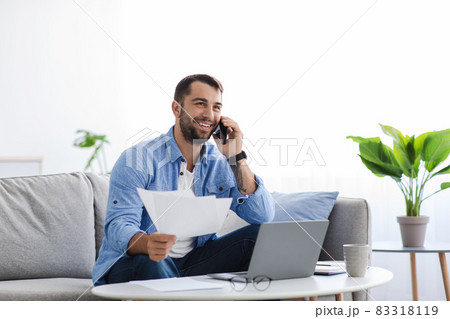 Busy smiling adult european guy sitting on sofa work at home with documents, talking with client by phone Busy smiling adult european guy sitting on sofa work at home with documents, talking with client by phone 83318119