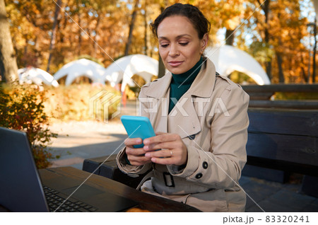 Confident business woman, young freelancer, texting messages on mobile phone while working remotely, sitting on terrace of a wooden cafe in an oak grove on beautiful autumn warm sunny day Confident business woman, young freelancer, texting messages on mobile phone while working remotely, sitting on terrace of a wooden cafe in an oak grove on beautiful autumn warm sunny day 83320241