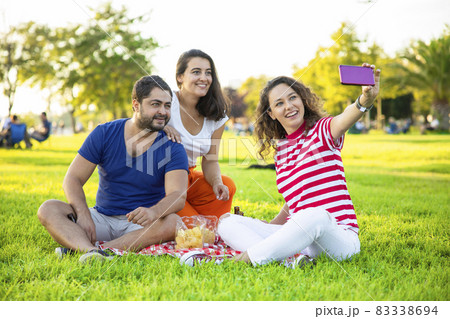 Three friends sitting on the grass and taking a selfie Three friends sitting on the grass and taking a selfie 83338694
