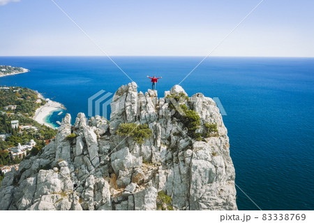 Aerial view of man tourist in red jacket standing on the rock top of cat mountain and raising his hands in front of landscape of Simeiz cosatline. Crimea 83338769