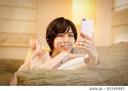 ソファに寝転んでスマホを見る若い女性 83340885