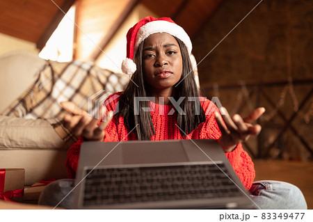Upset black lady in Santa hat having virtual call on laptop during Christmas, feeling sad on winter holidays at home Upset black lady in Santa hat having virtual call on laptop during Christmas, feeling sad on winter holidays at home 83349477