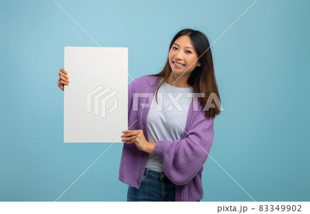 Cheerful asian lady holding blank board for advertisement or text, standing over blue studio background 83349902