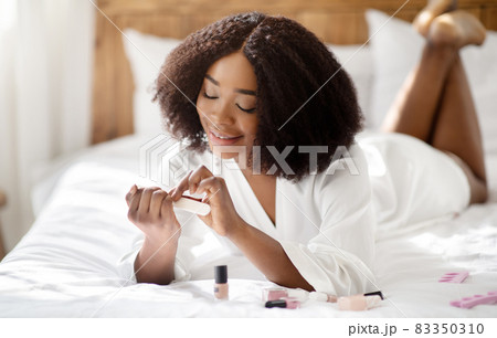 Portrait of lovely black woman filing her nails, making manicure on bed at home. Spa procedures, beauty treatments Portrait of lovely black woman filing her nails, making manicure on bed at home. Spa procedures, beauty treatments 83350310