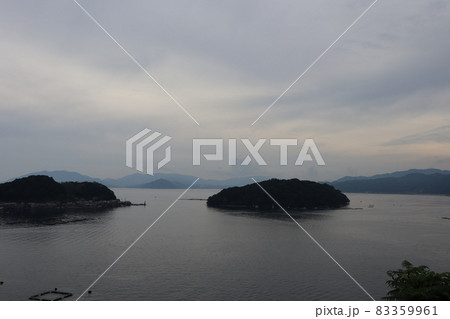 高台から見た養殖いかだと舟屋や民家のある町の半島と海と霞んで見える山々と曇り空の風景 83359961