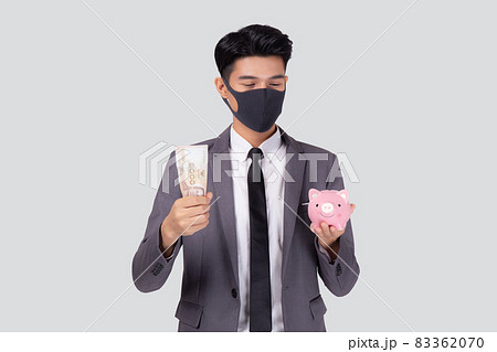 Young asian businessman in face mask holding money Thai and piggy bank isolated. Young asian businessman in face mask holding money Thai and piggy bank isolated. 83362070