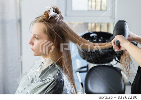 Close up of hairdressers hands drying long blond hair with blow dryer and round brush Close up of hairdressers hands drying long blond hair with blow dryer and round brush 83362280