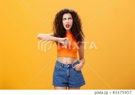 Party for losers I am out. Dissatisfied attractive popular girl with curly hairstyle in red lipstick and cropped top rolling eyes up from irritation and dislike showing thumb down over orange wall Party for losers I am out. Dissatisfied attractive popular girl with curly hairstyle in red lipstick and cropped top rolling eyes up from irritation and dislike showing thumb down over orange wall 83375957