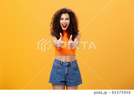 Charismatic ambitious and outgoing charming caucasian woman with curly hairstyle and red lipstick showing thumbs up gesture in like or approval smiling joyfully being supportive over orange wall 83375959