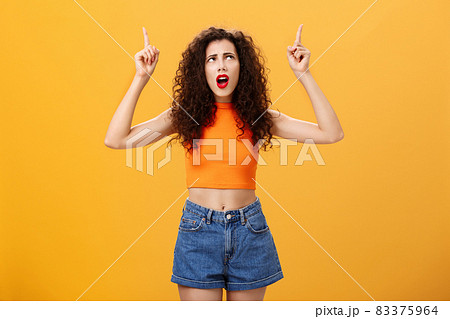 Silly and moody european party girl with curly hairstyle in red lipstic and stylish orange cropped top complaining on bad weather raising hands pointing and looking up with displeased unhappy face Silly and moody european party girl with curly hairstyle in red lipstic and stylish orange cropped top complaining on bad weather raising hands pointing and looking up with displeased unhappy face 83375964