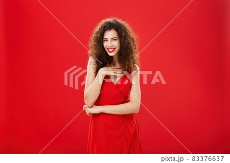 Woman receiving compliments attending luxury party in evening dress gently touching breast and flirting with sensual smile standing over red wall with curly hairstyle chuckling answering on guy flirt Woman receiving compliments attending luxury party in evening dress gently touching breast and flirting with sensual smile standing over red wall with curly hairstyle chuckling answering on guy flirt 83376637