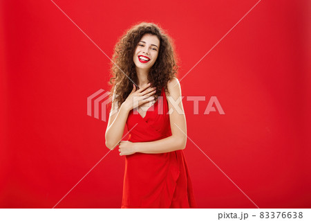 Portrait of elegant pleased and grateful charming caucasian woman with curly hairstyle holding palm on chest smiling and laughing amused posing in elegant evening dress over red background Portrait of elegant pleased and grateful charming caucasian woman with curly hairstyle holding palm on chest smiling and laughing amused posing in elegant evening dress over red background 83376638
