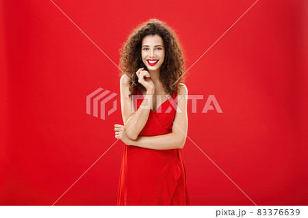 Passionate charming european woman over red background in elegant dress with curly hairstyle smiling cute, feminine playing with hair strand standing timid and silly, talking to person she admires 83376639