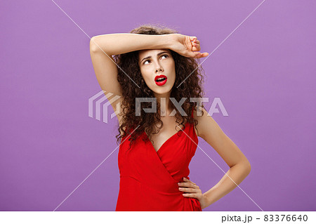 Troubled and tired good-looking european woman with curly hairstyle in red evening dress wiping sweat of forehead looking at upper right corner perplexed and exhausted over purple background Troubled and tired good-looking european woman with curly hairstyle in red evening dress wiping sweat of forehead looking at upper right corner perplexed and exhausted over purple background 83376640
