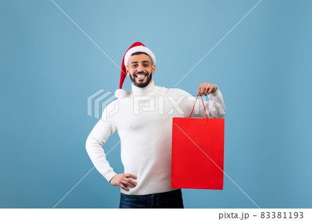 Happy arab man wearing Santa hat and holding shopping bag with Christmas gift over blue background, studio shot 83381193