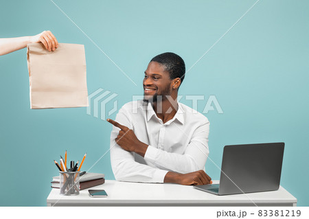 African american company worker receiving food delivery from courier and pointing on paper bag, blue background African american company worker receiving food delivery from courier and pointing on paper bag, blue background 83381219