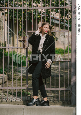 Close-up photo of young beautiful woman in black fur coat, jeanse and shoes posing on street Close-up photo of young beautiful woman in black fur coat, jeanse and shoes posing on street 83381957