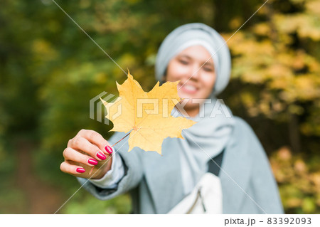 Autumn muslim woman holding maple leaf outdoors in sunny day. Beautiful arab wearing hijab girl in fall park 83392093
