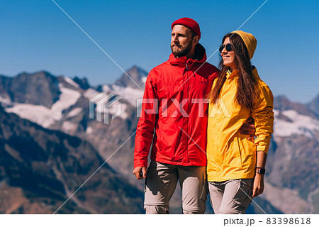 A traveling couple in hiking gear against the background of mountains. Two tourists on the top of the mountain. A man and a woman in the mountains. Two travelers in colorful jackets, close-up. 83398618