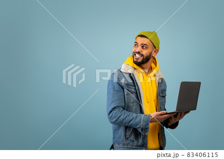 Remote job. Happy young arab guy with laptop computer looking aside at free space, standing on blue background 83406115