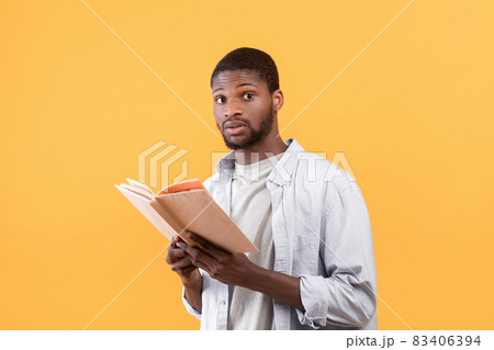 Shocked black male student holding textbook, looking at camera with shock over yellow studio background 83406394