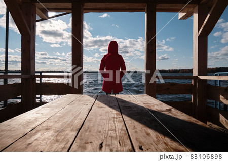 A woman in a Red Jacket Sits at a Table on a Wooden Pier and Looks at the Water, Clouds and Trees with Yellow Foliage. Autumn Landscape. 83406898