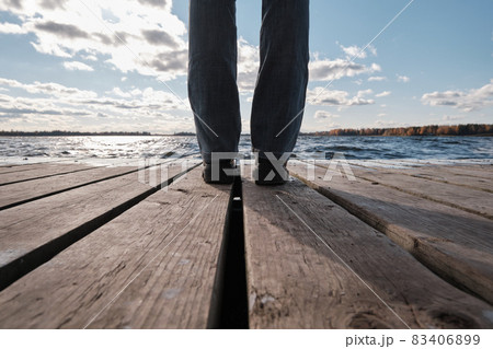 A woman in a Red Jacket Sits at a Table on a Wooden Pier and Looks at the Water, Clouds and Trees with Yellow Foliage. Autumn Landscape. A woman in a Red Jacket Sits at a Table on a Wooden Pier and Looks at the Water, Clouds and Trees with Yellow Foliage. Autumn Landscape. 83406899